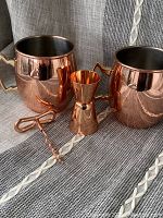 Photo showing two copper Moscow Mule mugs, a copper jigger, and a copper corkscrew arranged on fabric surface.