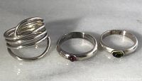 Three sterling silver rings displayed together on a flat surface: wide multi-band ring, and two thin bands each with a small stone (red and green).