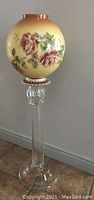 Full view of the tall glass lamp post with clear glass stem and base, topped with a large spherical glass shade painted with pink roses on a cream background.