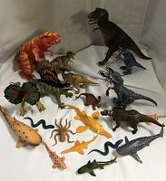 Photo showing the majority of the dinosaur and prehistoric creature figures arranged on a white backdrop. Includes large orange T-Rex, Dimetrodon with sail back, several small sea creature figures like sharks, eels, and an octopus.