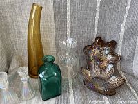 Seven-piece vintage glassware lot on light textured fabric backdrop, showing amber tall vase, green bottle vase, clear Scandinavian style ruffled top vase, two iridescent decanters, and two iridescent flower-shaped dishes.
