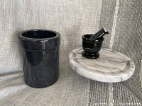 Full set displayed together on fabric backdrop showing black marble utensil holder on left and white marble cheese board with black mortar and pestle on right.