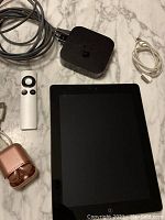 View of all items in the lot on marble floor: iPad 2, Apple TV with remote and cables, rose gold wireless earbuds in case, charging cable.