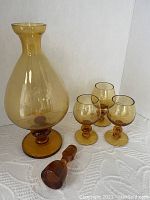 Photo of amber glass decanter without stopper and three matching amber stemmed glasses on white textured surface.