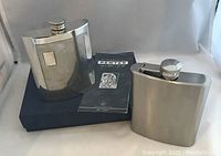 Two drinking flasks side by side on a black box; one stainless steel with polished and textured design, the other smooth stainless steel; includes a small pamphlet.