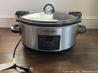 Full exterior view of Crock-Pot showing stainless steel body, clear glass lid with locking flaps, digital control panel, and power cord.