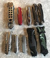 Top-down view of all 9 camping utility blades and multi-tools arranged on surface showing variety of handles and attachments.