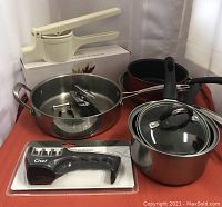 Photo shows 3 pots (two stainless steel, one black), kitchen gadgets including a white handheld unit, and a packaged black blade sharpener.