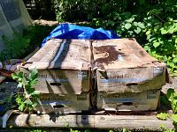 Photo showing four worn cardboard boxes of decorative cultured stone on a wooden pallet with natural surroundings and blue tarp partially visible.