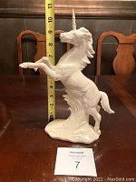 Full view of the white ceramic unicorn figurine showing its size against a measuring tape and background chairs.