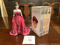 Frontal view of the Royal Worcester Audrey Hepburn figurine in pink gown next to the boxed Audrey Hepburn VHS tape collection