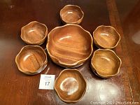 Seven wooden bowls arranged on table showing the large bowl in center and six smaller matching bowls around it, wood grain and scalloped edges visible.