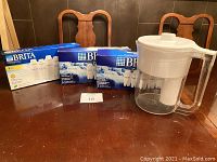 Brita water filter pitcher and three boxes of replacement filters arranged on wood table with chairs in background