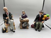 Front view of the three Royal Doulton figurines: seated man with bottle, seated woman at desk, seated man with balloons.