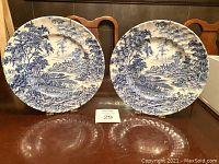 Two matching blue and white Ridgway ironstone plates with detailed pastoral scene, scalloped edges, displayed upright on a wood table.