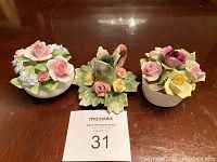 Three vintage porcelain flower arrangements on wooden table with auction lot number card