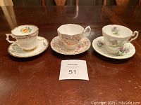 Three vintage teacup and saucer sets displayed on a dark wooden table, showing overall lot layout and detail from front angle.