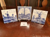 Front view of three Delft blue ceramic tiles displayed on stands, showing intricate traditional Dutch scenes of windmills and Amsterdam landmark.