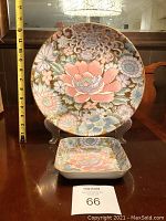 Front view of 12 inch round plate and smaller square trinket plate on display stands, showing detailed floral pattern with pink, lavender, green, and gold accents