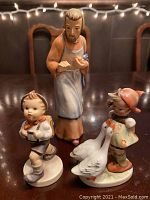 Three porcelain Hummel figurines shown on table with string lights in background.