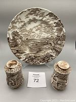Round vegetable plate decorated with a brown transferware countryside village scene, displayed upright on a clear stand behind two cylindrical salt and pepper shakers with matching brown village transfer patterns.