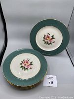 Group photo of 8 dinner plates with green rim, gold edge and pink floral center stacked, and one plate displayed upright.
