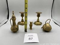 Five antique brass items on a white background including two candleholders, one tall ornate pitcher, one teapot-style jug, and one small jug. A tape measure shows height around 7 inches for the pitcher.