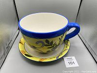 Photo showing large handpainted ceramic cup with blue rim and handle, sitting on matching saucer with handpainted floral design.