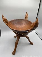 Front view of antique wooden bowl on carved wooden stand with human head carvings on rim and leg-shaped feet.