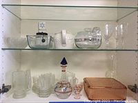 Shelved glassware and kitchen items including Pyrex coffee pot, glass pitcher, vintage grinder, decanter, lidded serving bowl, and glassware.