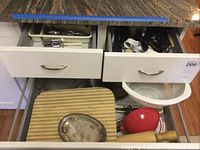 Two open drawers showing stainless steel flatware and kitchen utensils including cutting board, wooden rolling pin, two plastic strainers, and silver plated dish visible below drawer