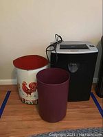 Photo showing the Fellowes paper shredder with cord, a maroon plastic trash bin, and a white metal trash bin with rooster design.