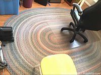 Full view of oval braided area rug placed on hardwood floor under chair and nearby plastic container.