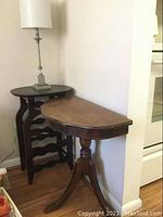 Shows the dark wooden wine bottle holder side table with wavy bottle rack and flat round top, a demi lune half-moon shaped side table with worn wood finish, and a lamp with light fabric shade on top of wine holder table.