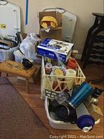 Photo showing the small wooden stool near the door with various household items in bins including ceramic figurines, boxed label maker, mugs, and candles on the floor.