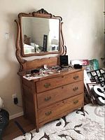 Front and angled view of antique wooden dresser with three drawers and attached ornate mirror frame showing carved wood detail and brass drawer handles. Scratches visible on dresser surface.