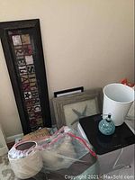 Photo showing various household items grouped including pillows in plastic bag, framed art leaning against wall, white trash bin, a glass soap dispenser, and black digital bathroom scale on top of wooden towel warmer.