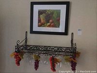 Photo of metal wine rack mounted on wall showing ornate design, hanging green and red faux grapes, and hanging wine glasses beneath rack, with framed fruit print above rack.
