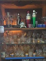 Photo showcasing assorted glassware including shot glasses, cordial glasses, salt cellars, cocktail shakers in silver and green metal, a glass decanter, and a small decorative turtle figurine on a wooden shelf.
