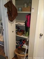 Open cabinet with wicker laundry basket, colorful fabric shoulder bag, towels, pillow cases, and plastic jars