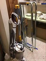 Shark professional vacuum cleaner with attached hose and accessories, Hangaway clothes hanger, and metal cot frame folded, items shown in room with shelves and dishes in background.