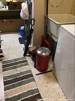 Striped runner rug on floor with adjacent household maintenance items including red trash can, blue bucket, black umbrella, and floor sweeper.