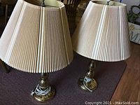 Pair of brass lamps with pleated cream-colored shades on carpeted floor, showing full lamps and cords coiled at base.