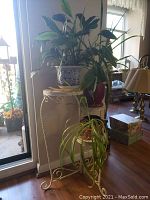 Three-tier metal plant stand in white with mosaic tile shelves holding three potted plants. The plants are different types, varying in leaf shape and size.