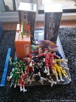 Multiple Power Rangers action figures in various colors lying on a flat surface next to the orange plastic lunchbox and two boxed Hess toy trucks.