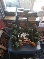 Overall display of boxed Victorian Village collectible buildings with loose pieces including miniature pine trees and plastic reindeer on a small table near a window.