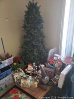 Full view of assembled 6ft artificial Christmas tree surrounded by assorted holiday decorations including gift boxes, assorted figurines, ornaments, linens, and storage containers scattered around the base.