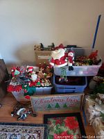 Photo of Christmas decorations and figurines on a wooden table, includes bins with ornaments and figurines, red bows, artificial greenery, Santa and Snowman plush figurines, and Christmas signage.