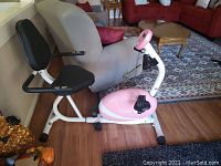 Side view of pink recumbent bike showing seat, pedals, and frame on hardwood floor.