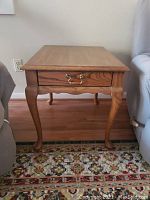 Front view of Mersman Waldron Comfort side table with single drawer and brass handle, medium oak finish, placed on hardwood floor near couch on patterned rug.